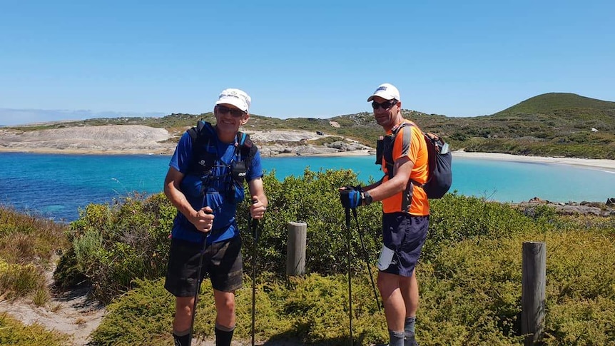 Two men in high-vis clothing and backpacks pose leaning on their walking poles on a bushy cliff overlook blue waters.