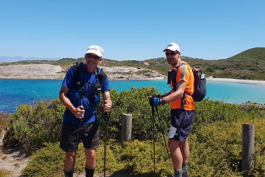 Two men in high-vis clothing and backpacks pose leaning on their walking poles on a bushy cliff overlook blue waters.