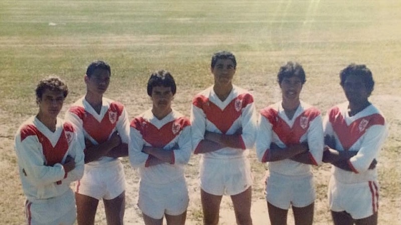 Six Indigenous teenagers in football clothing standing on a field with arms crossed.