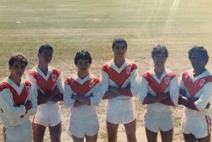 Six Indigenous teenagers in football clothing standing on a field with arms crossed.