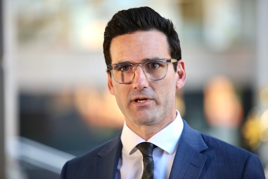 Chris Rodwell mid-speaking with a soft focused background. He wears a suit, tie and glasses.