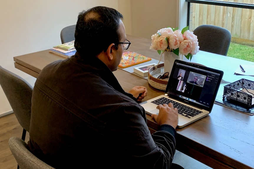 A man looks at a laptop computer seated at a table with a vase of flowers.