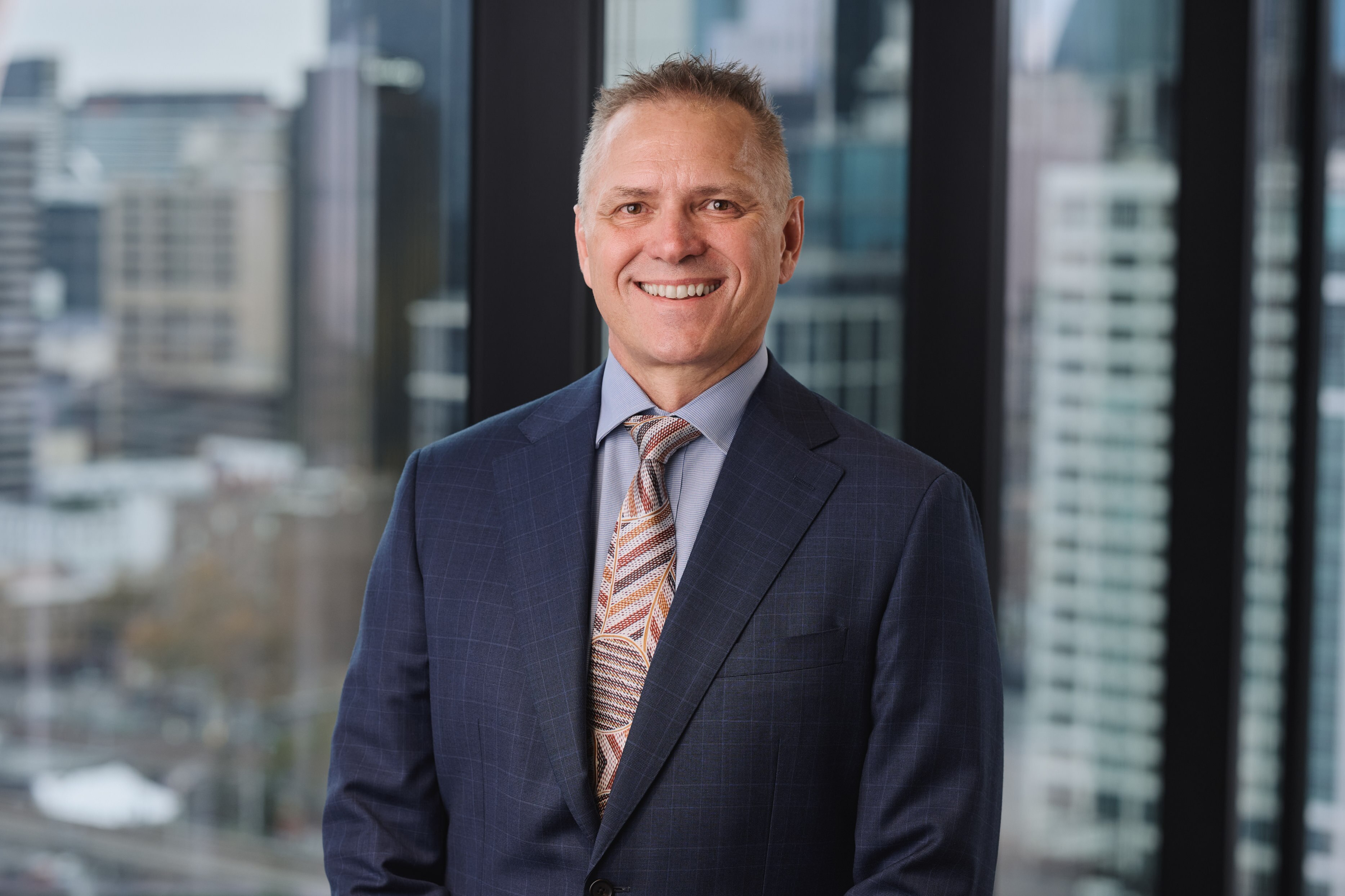 A smiling man in a suit, tie and close-cropped hair poses in front of a high-up window showing a cityscape.