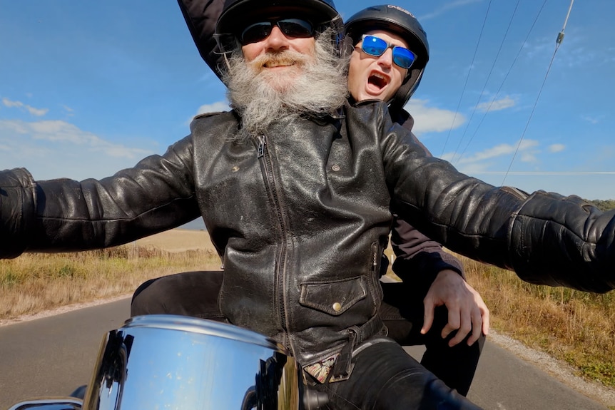 Two men on a motorbike on an open road, arms out stretched and shouting jubilantly. 