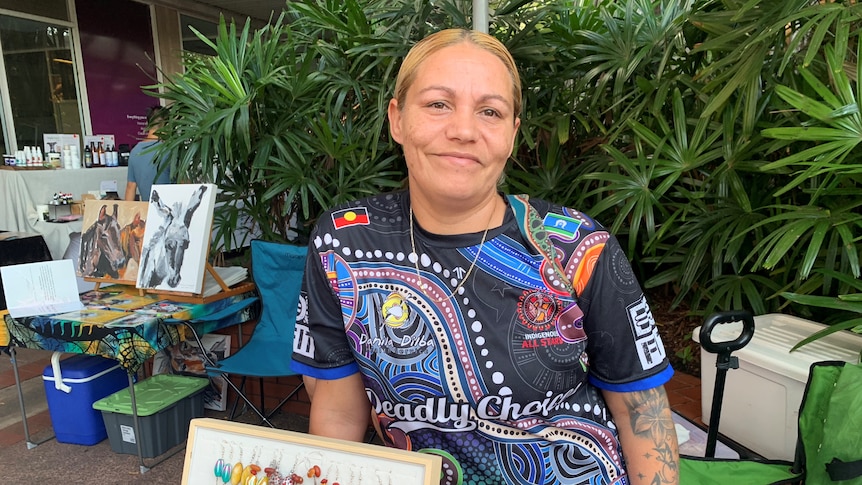 Woman with Indigenous t-shirt holds a piece of art up while standing in front of a bush
