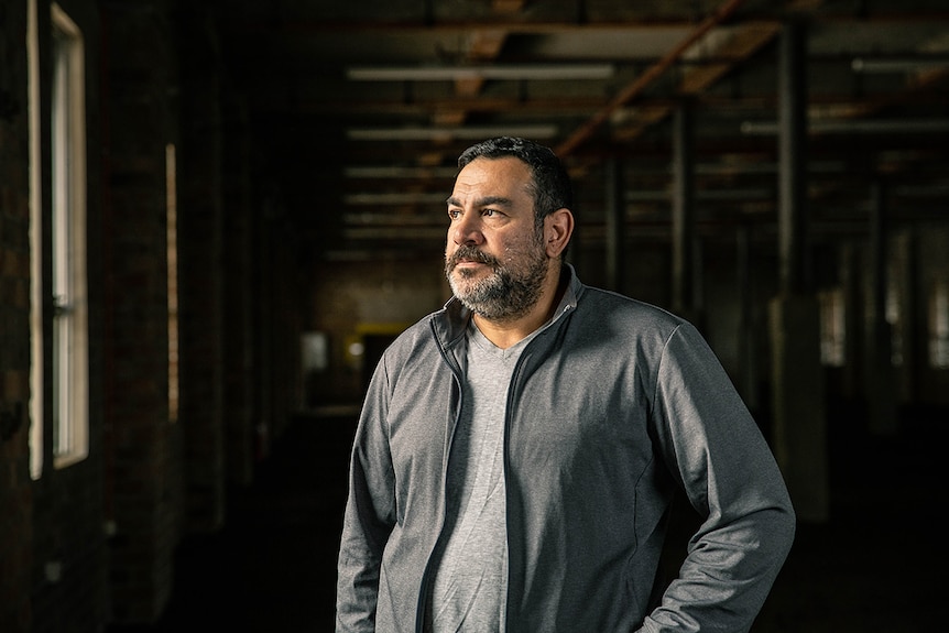 Colour photo of artist Khaled Sabsabi looking out the window in a dimly lit brick warehouse.