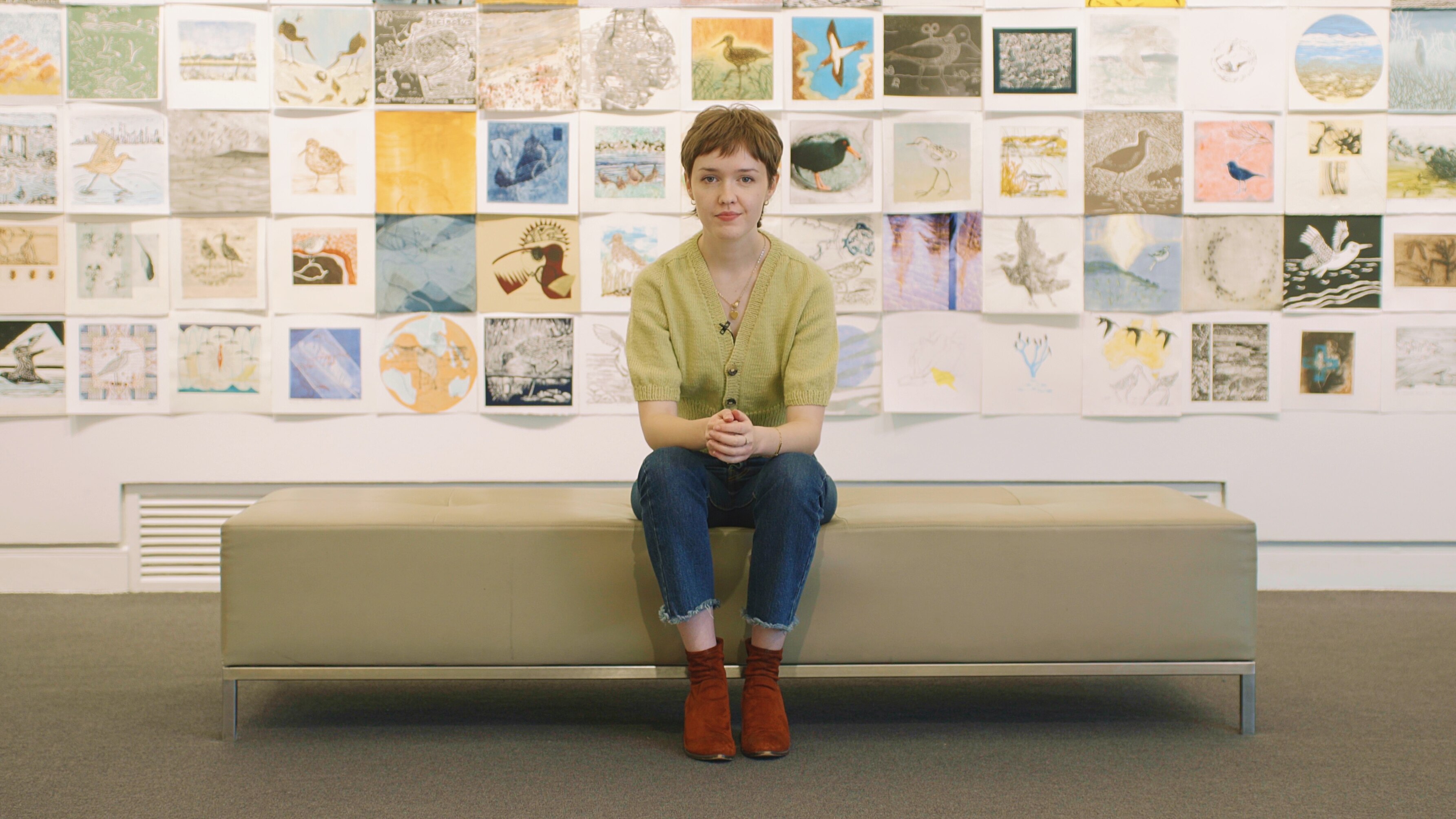 Mary McGillivray, a young woman with a short haircut, sitting in front of a wall of drawings at a gallery