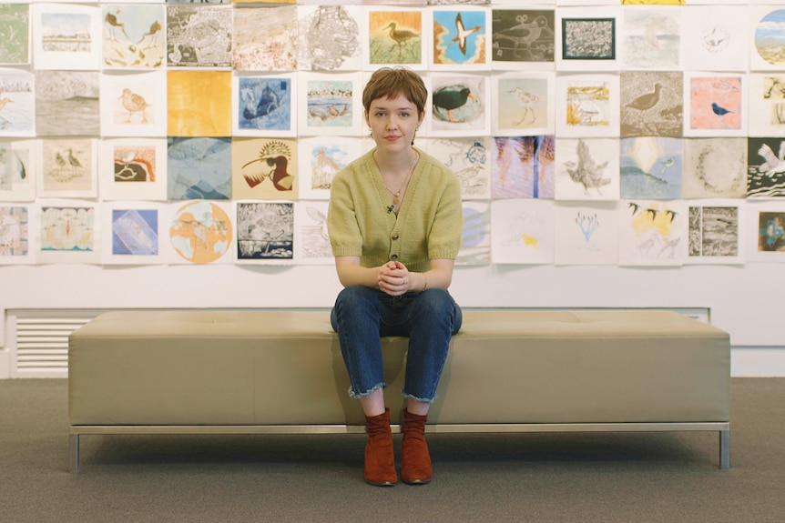 Mary McGillivray, a young woman with a short haircut, sits in front of a wall of drawings in a gallery