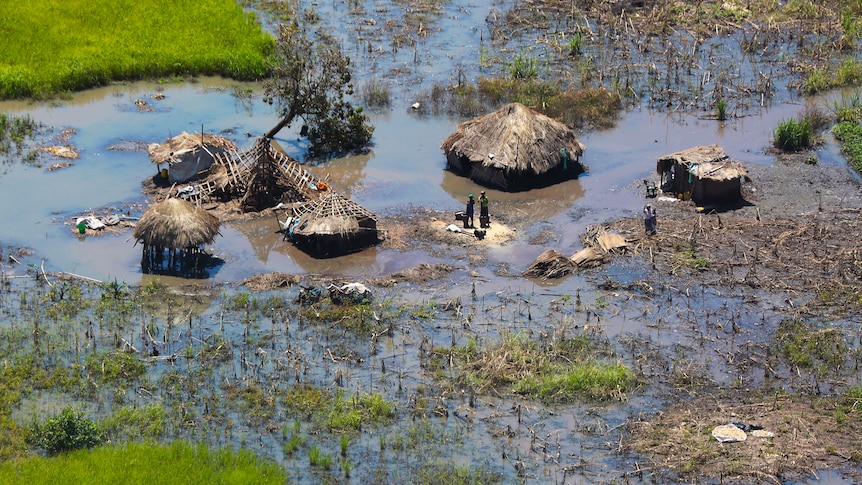 A flooded village area show people stranded around primitive huts on grassy plain