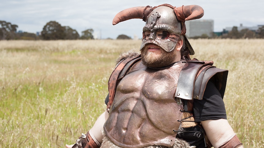 A young man with a beard stands in a viking costume in a field of long, dry grass.