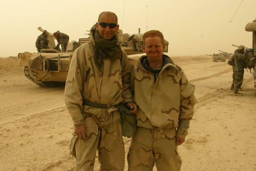 Two men in military gear stand in front of an army tank in Iraq in 2003.