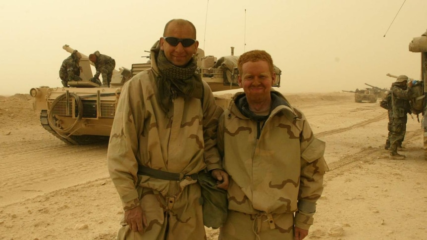 Two men in military gear stand in front of an army tank in Iraq in 2003.