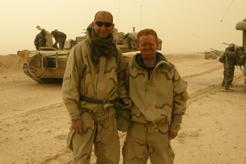 Two men in military gear stand in front of an army tank in Iraq in 2003.