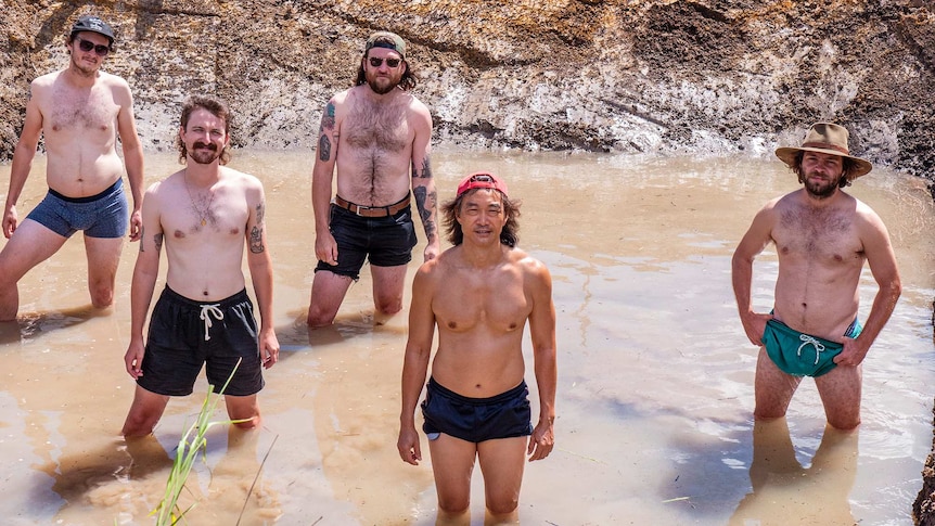 A group of shirtless men stand in a muddy dam like hole.