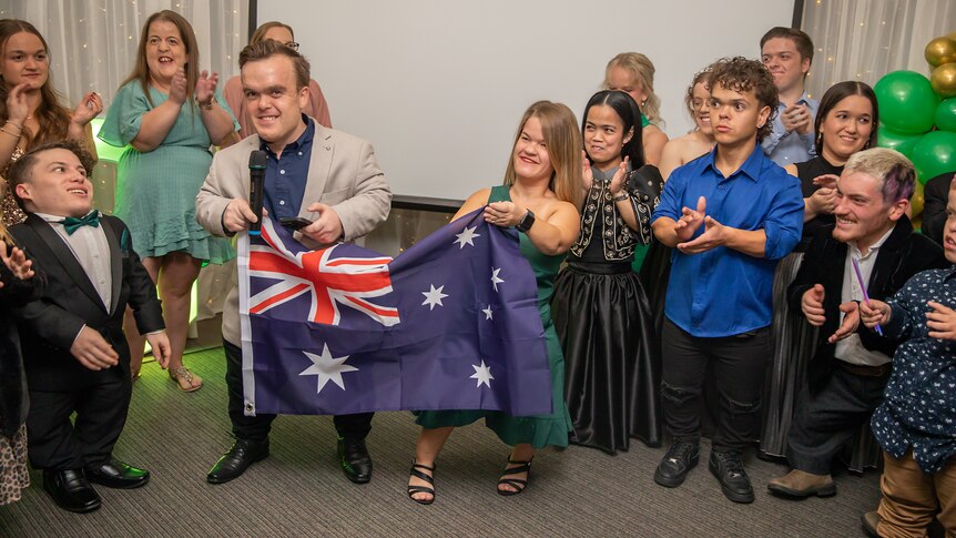 Samantha Lilly and Michael Spain hold an Australian flag. They are surrounded by clapping team members.