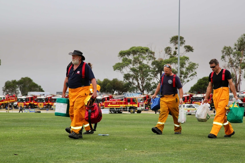 Three men and a woman in firefighting overalls walk across an oval with backpacks in hand.