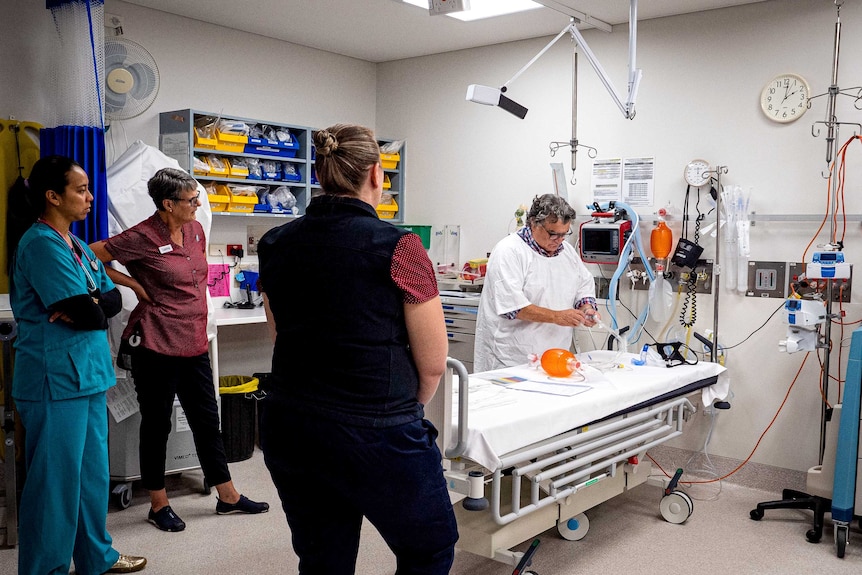 A group of staff stand around a hospital bed as they are shown how to use equipment that assists with breathing.