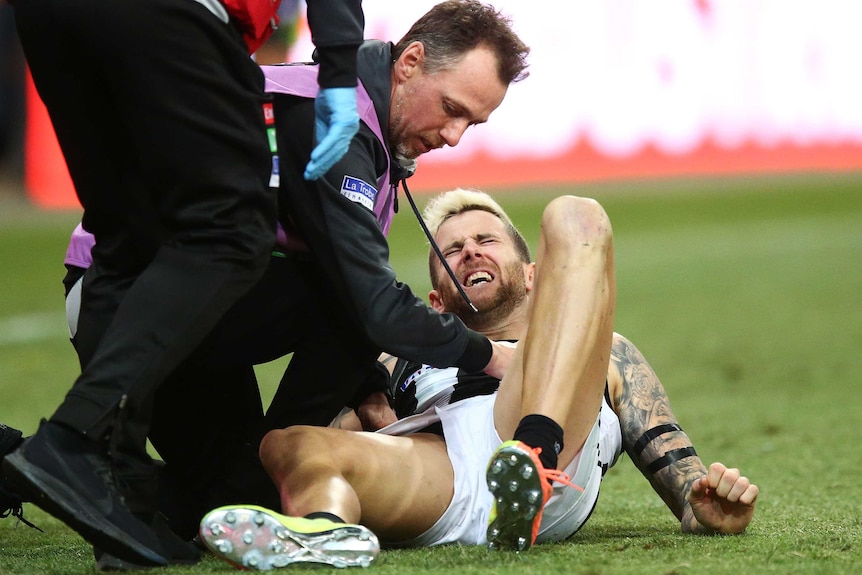 An AFL team staffer tests out an injured player as he lies on the ground, grimacing in pain.