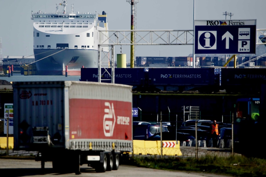 A truck trailer is parked at Zeebrugge port with a large ferry in the background.