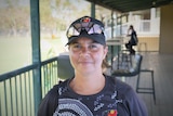 A woman in a cap and sport uniform, standing on the verandah of a country stadium and smiling.