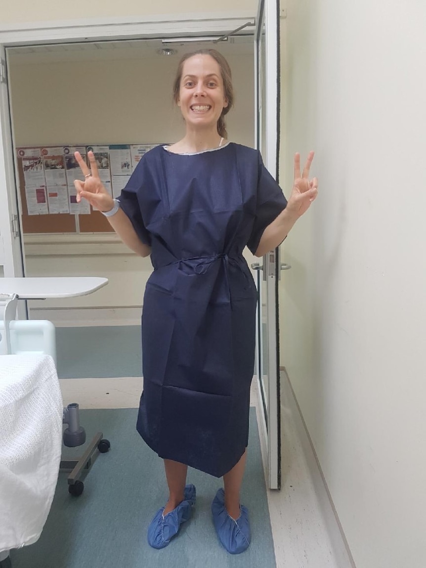 A woman wearing a navy hospital gown holding up both hands in 'peace' signs