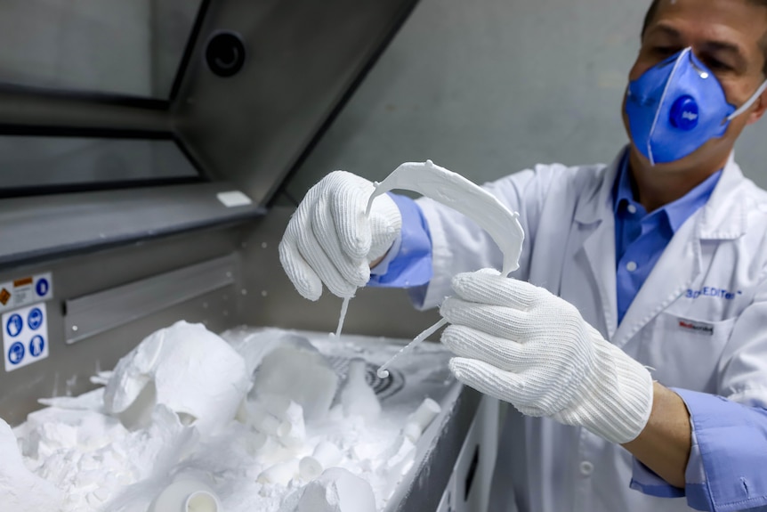 Man in white coat and blue mask holds up a white visor next to a 3D printer machine