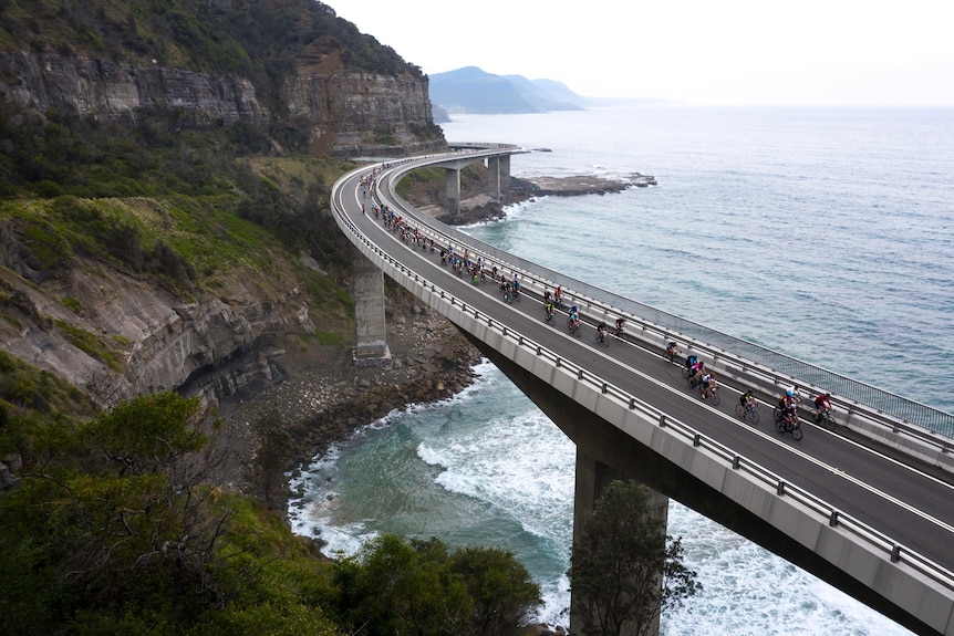 Bike riders ride along a bridge hugging the coast.