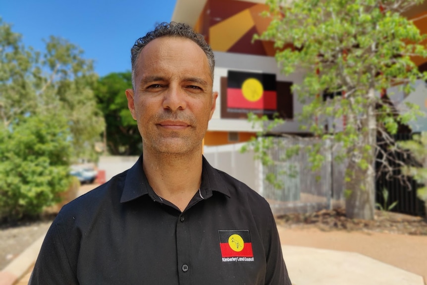 Tyronne Garstone smiling slightly in a portrait, taken in front of a building with the Aboriginal flag printed on the side.