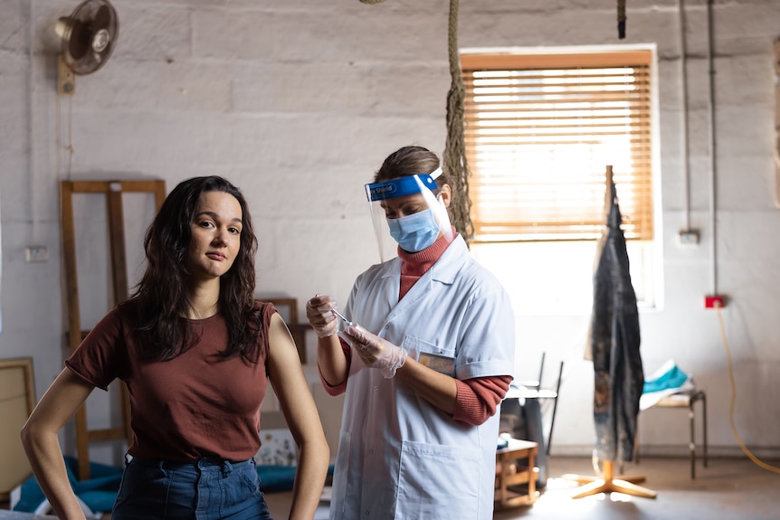 A young woman with long dark hair and a brown shirt sits looking up at a COVID-19 vaccination nurse in protective wear.