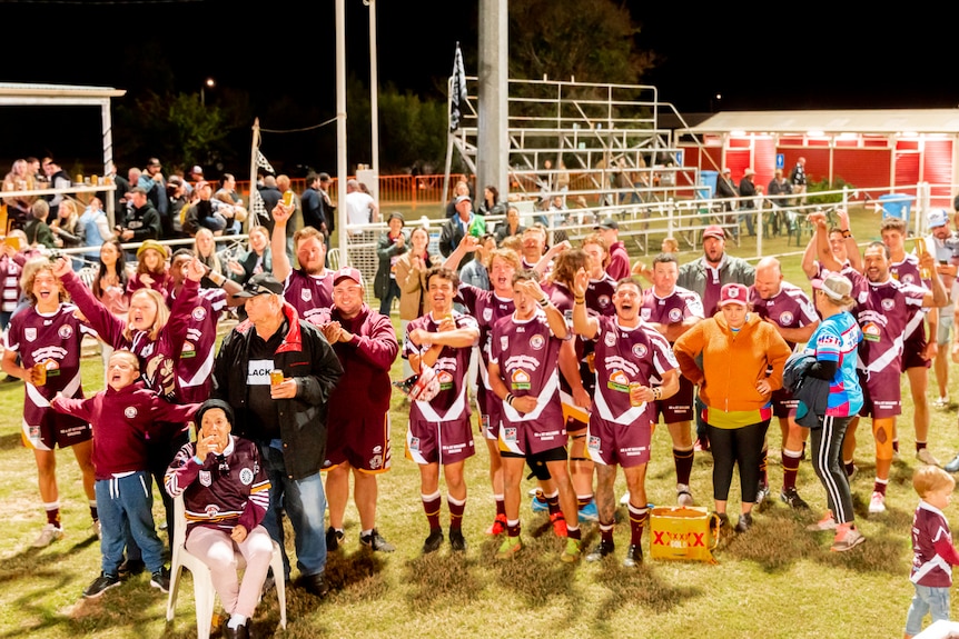 A big group of footy players from Barcaldine Western Queensland stand in maroon jerseys cheering after winning the grand final