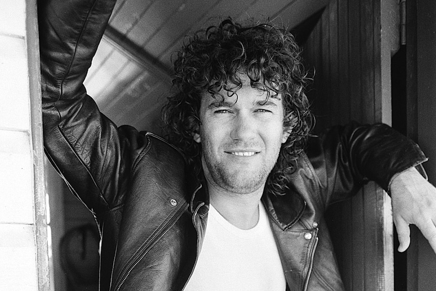 Jimmy Barnes in a leather jacket is leaning in a doorway with his arms on the sides of the doorway