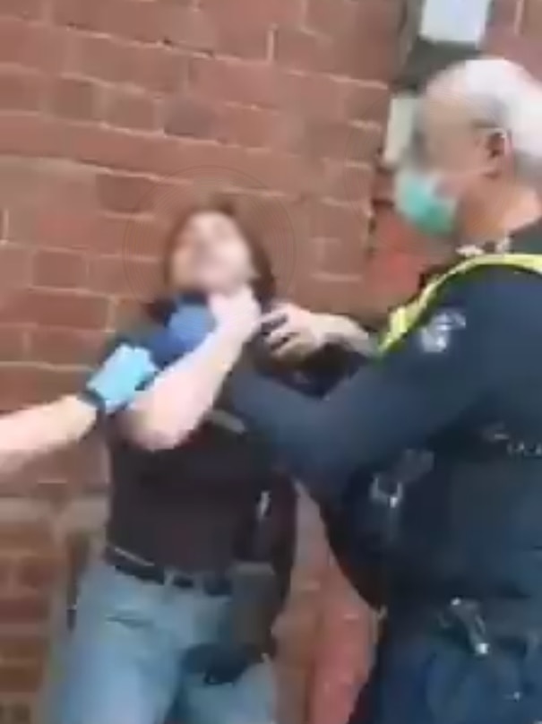 A Victoria Police officer wearing a blue surgical mask appears to be holding a woman without a face covering by the throat.