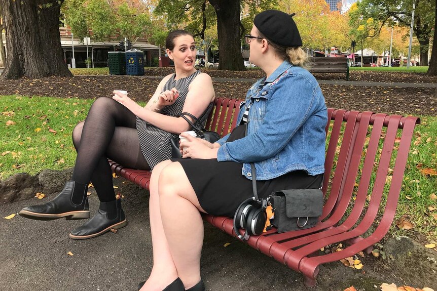 Two women sit on a park bench smiling at one another.