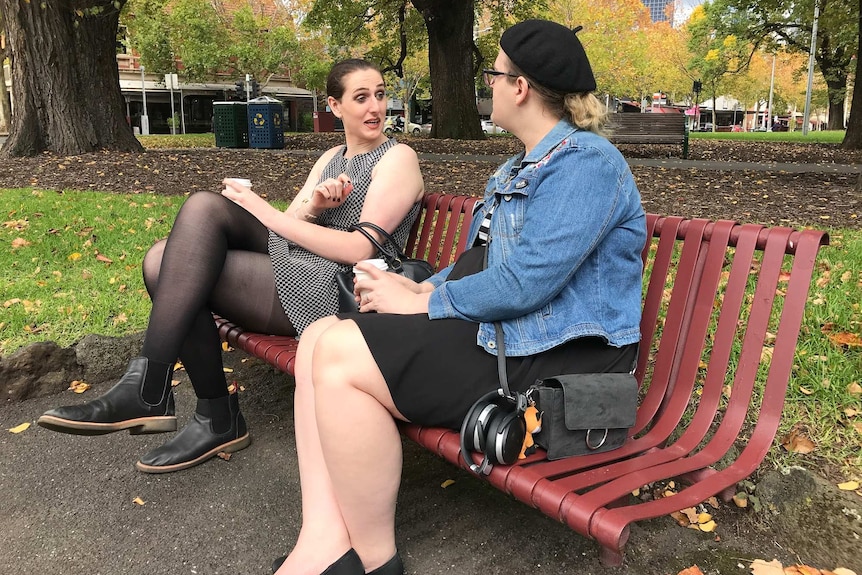 Two women sit on a park bench smiling at one another.