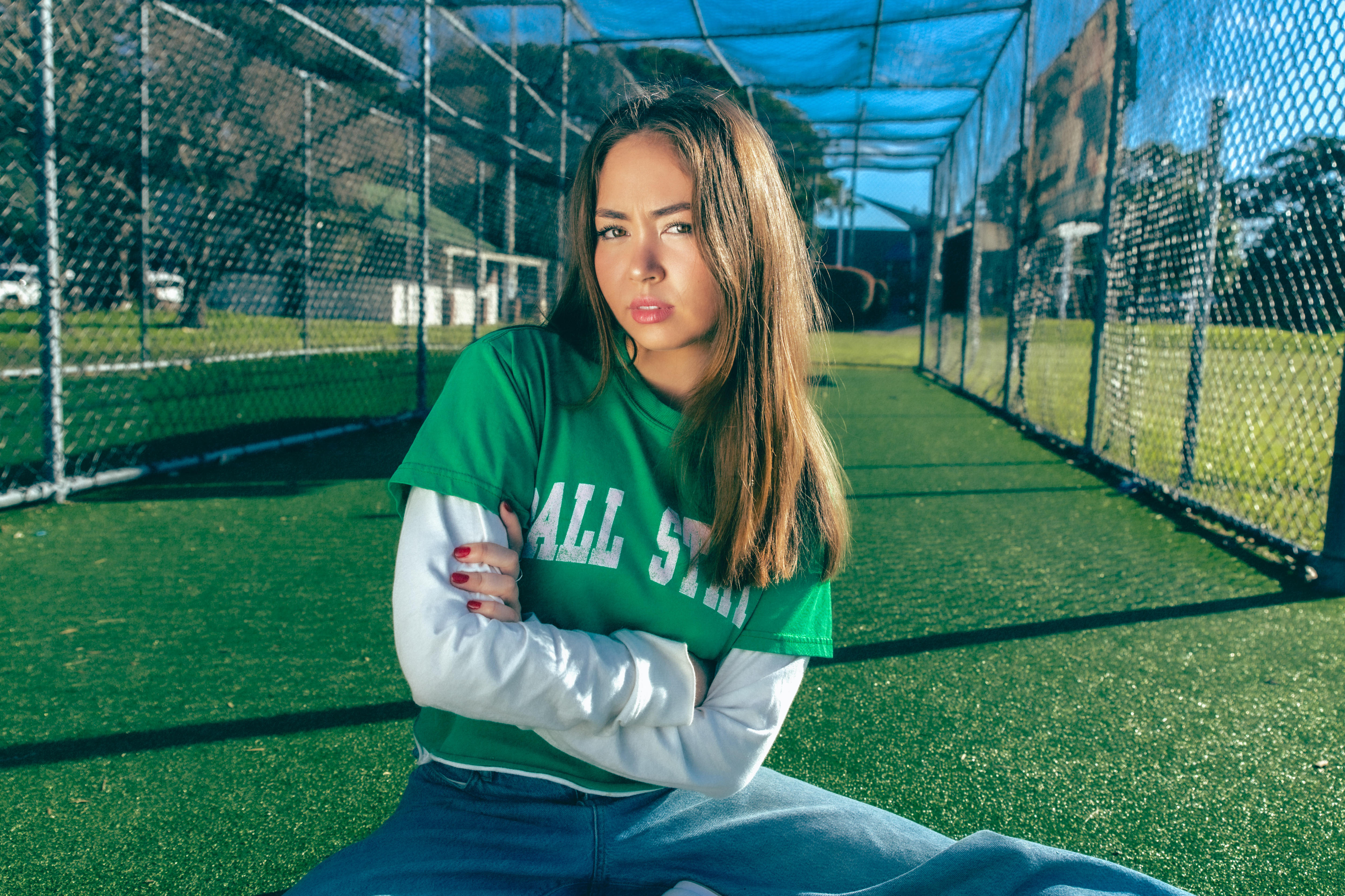 A woman in a green tshirt with a white longsleeve shirt underneath sits in a green astroturf cricket net with her arms crossed