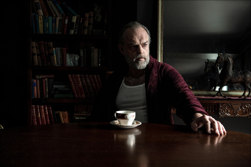 A film still of Hugo Weaving in a nightgown, sitting at a table with a cup of coffee in front of him.