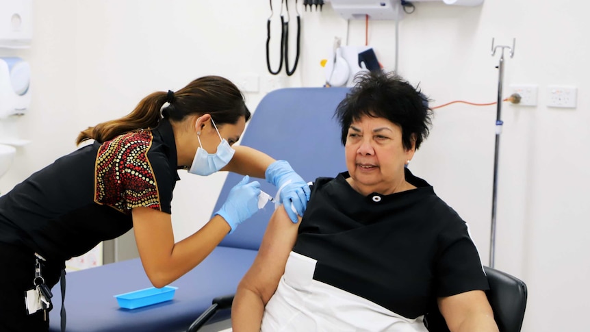 Dawn Casey sits in a chair as a young health worker delivers her first dose of vaccination against COVID-19.