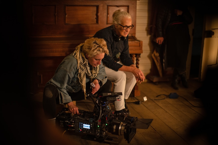 A 30-something woman holding a large video camera crouches beside a seated 60-something woman in a mid-century style room