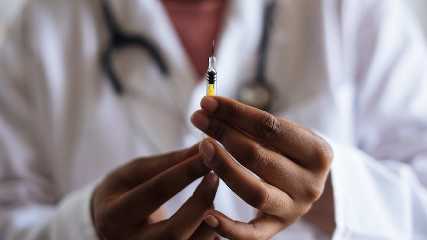 Hands holding a vaccine with a blurred figure of a person in a white coat in the background