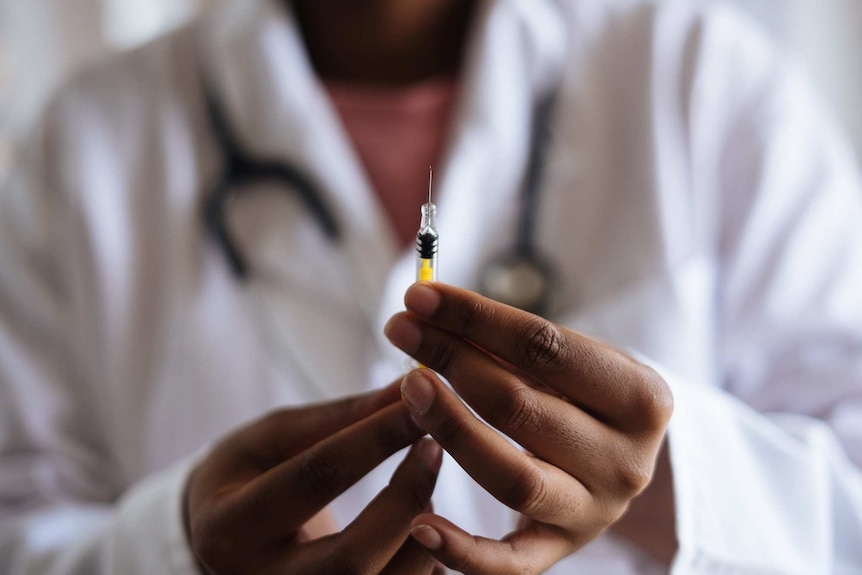 Hands holding a vaccine with a blurred figure of a person in a white coat in the background