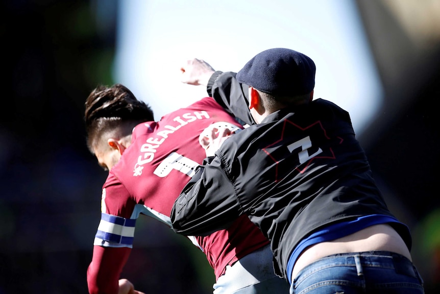 A man wearing a navy flat cap and black jacket punches Jack Grealish from behind in the head