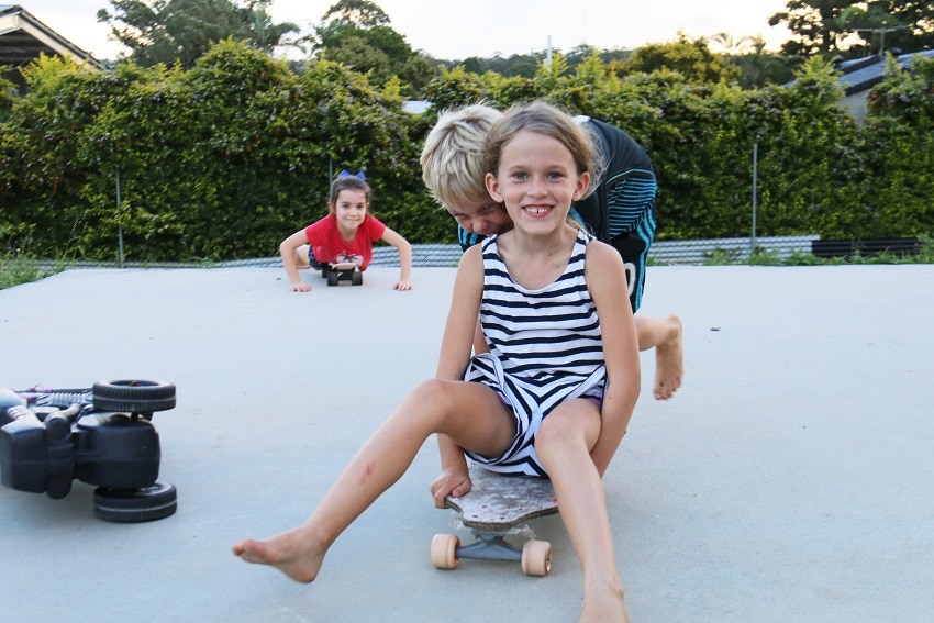 Mayla sitting on her skateboard pushed by one of her friends.