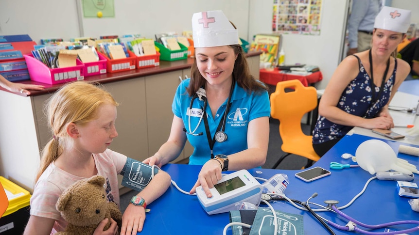 A woman shows a young girl how she checks blood pressure with a machine while the girl holds her teddy bear.