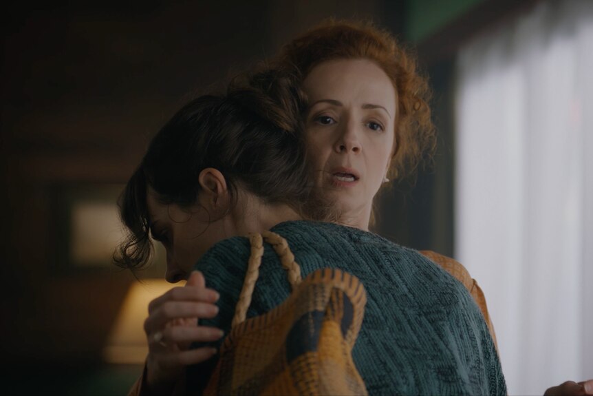 White woman woman with brown hair wearing blue cardigan embraces white woman with fiery red curly hair, who looks shocked.