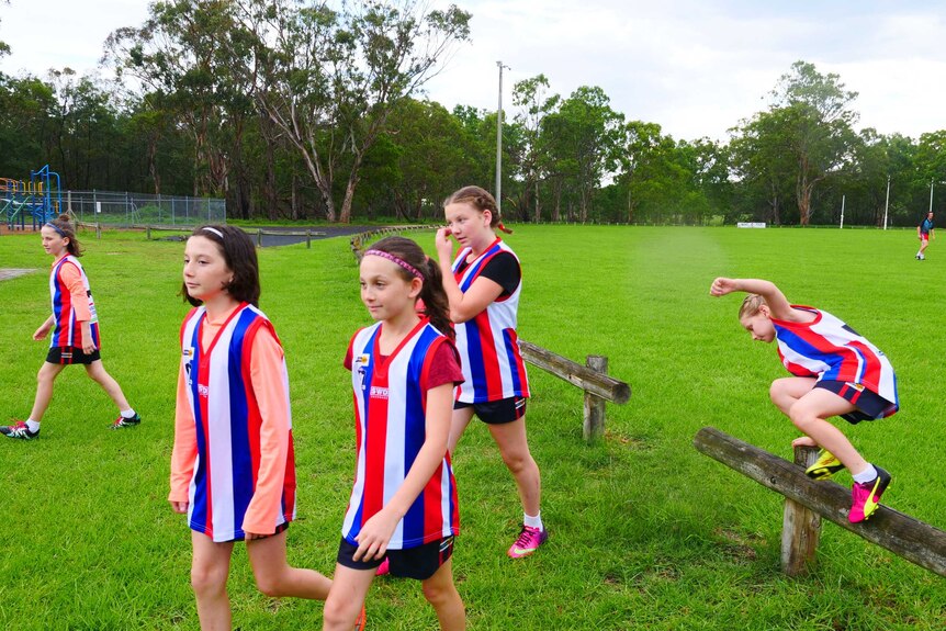 Five sisters jump a fence at footy oval, wearing red, blue and white striped football jumper.