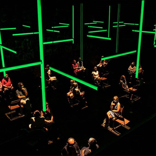 Wide shot of dozens of people wearing headphones sitting separated in a dark room while green-coloured bars of light hang above