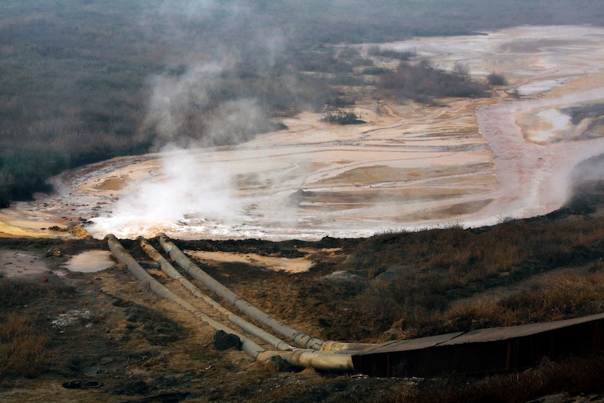 You look at a brown lake with black vegetation surrounding it as pipes pump mining waste into it, with steam rising from water.