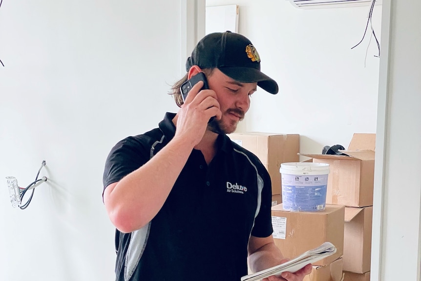 A man wearing a cap and a navy work polo has a phone to his ear, looking at a notepad. 