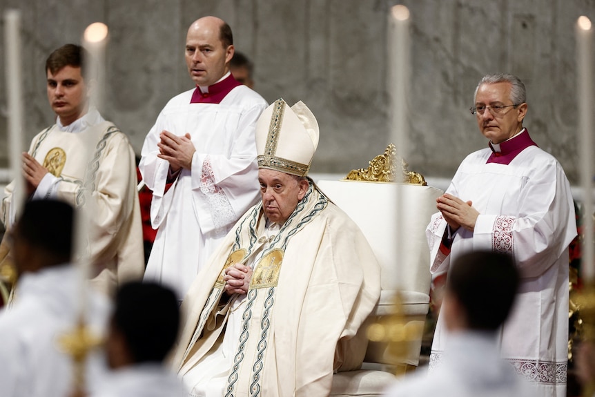 Pope Francis sits on a gold chair in robes surrounded by other priests. 