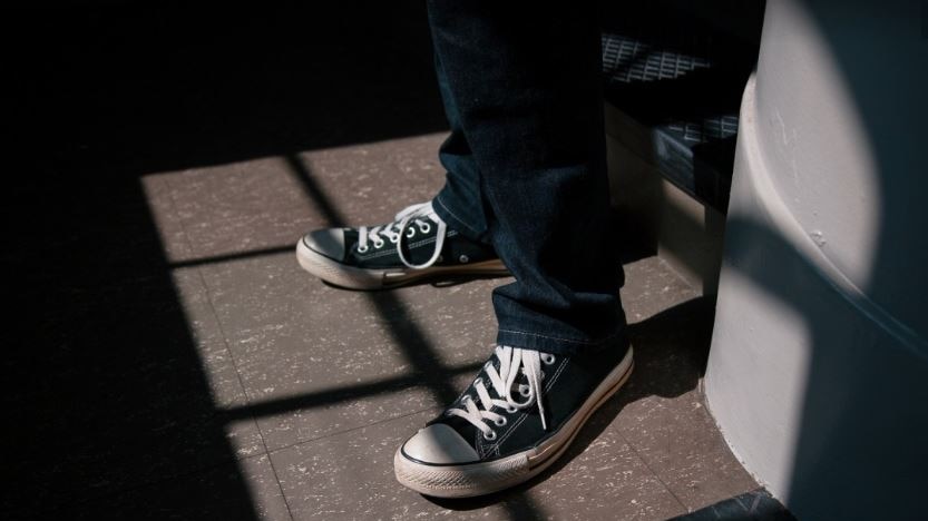 The bottom of a teenager's legs and his feet show as the boy stand at a window, wearing jeans and conversers.